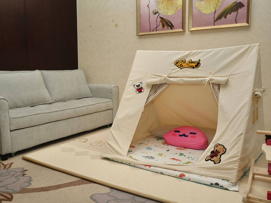 Une pièce avec un canapé gris, un petit espace de jeu aménagé comme une tente avec un coussin rose, et un tapis à motifs floraux.
