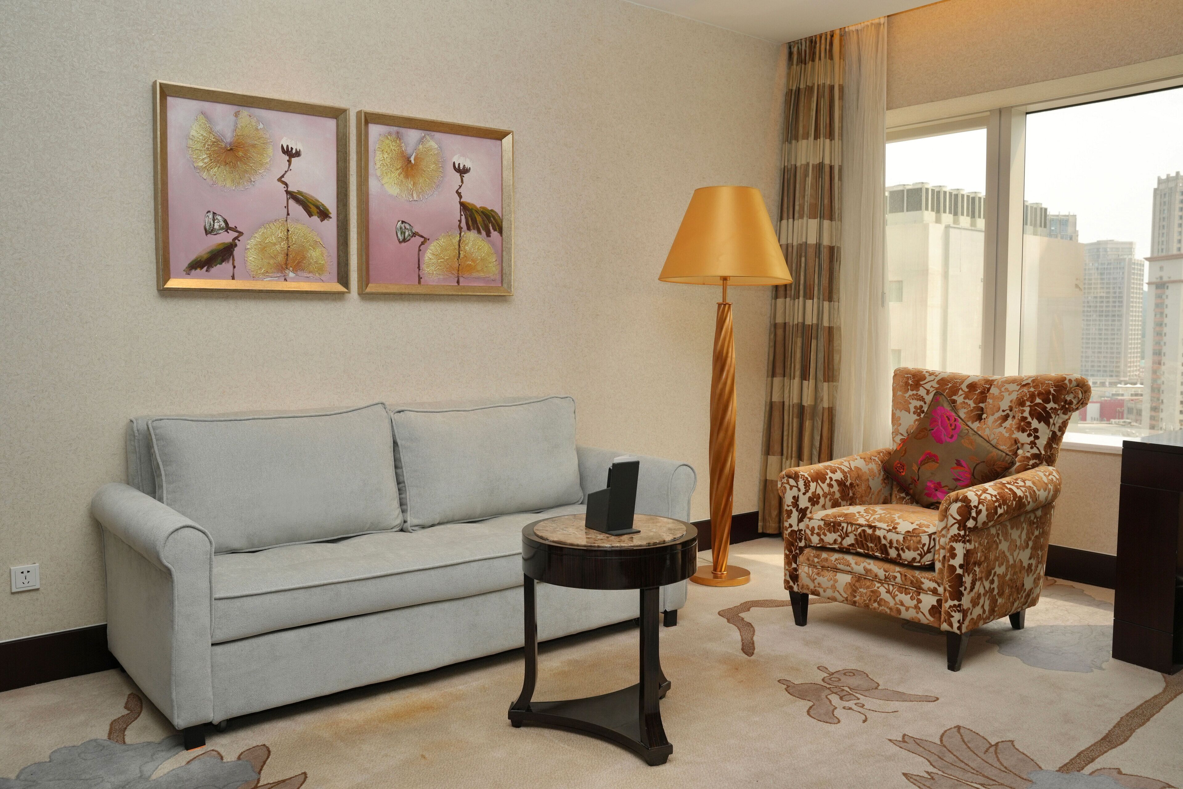 A living room with a light blue sofa, a patterned armchair, a small round table with a lamp, and two framed artworks on the wall.