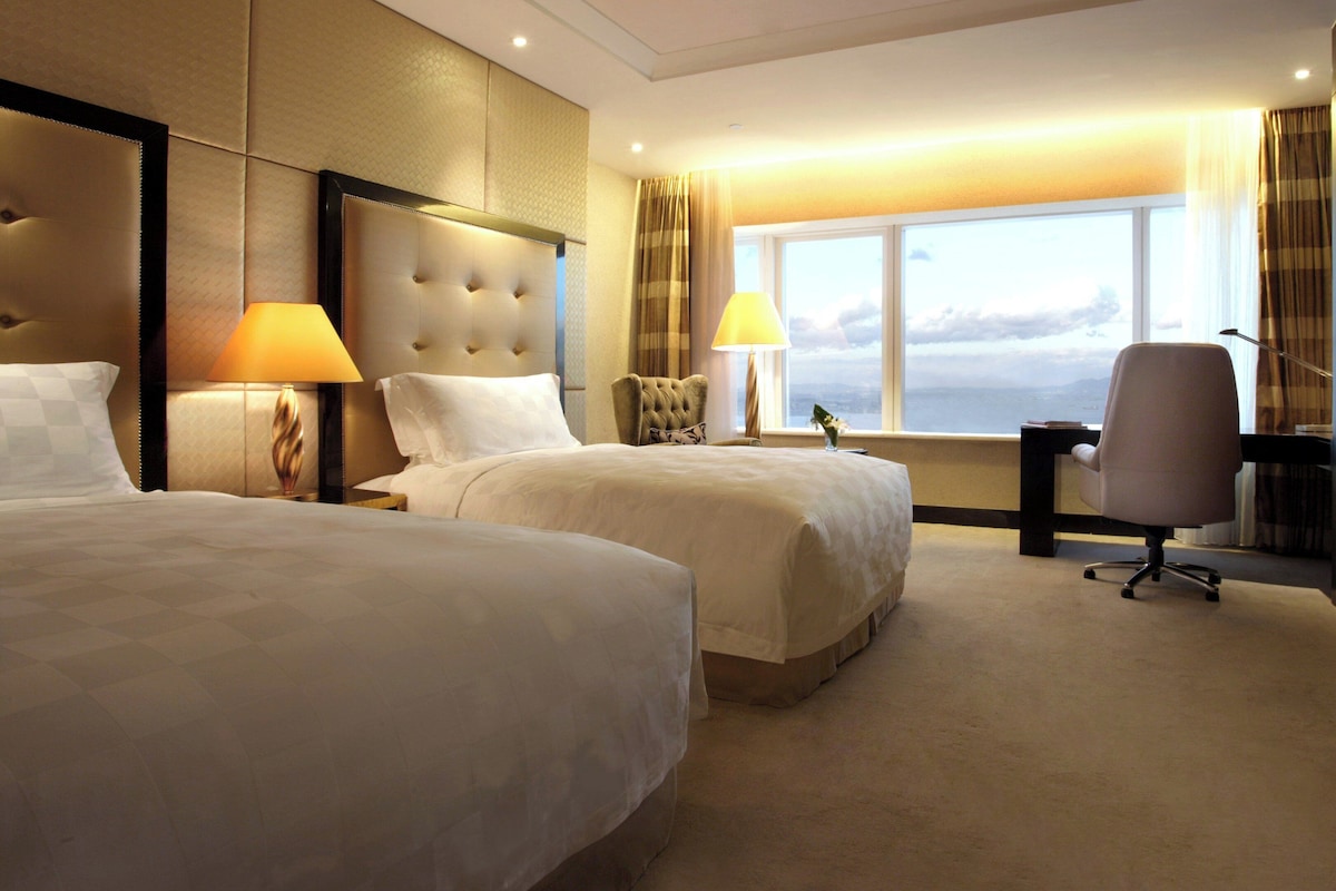 A hotel room with two beds, a desk, a chair, a TV, and a large window with a view.