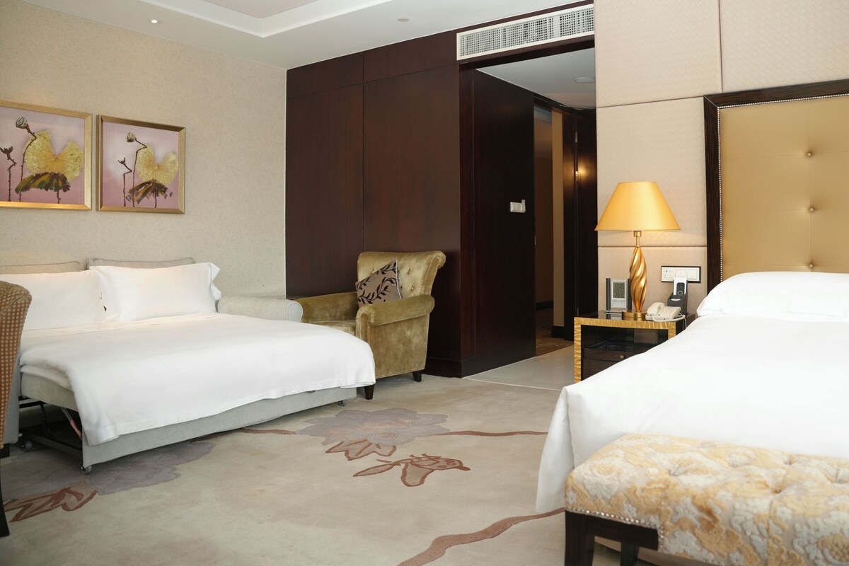 A hotel room with two beds, a chair, a lamp, and artwork on the wall.