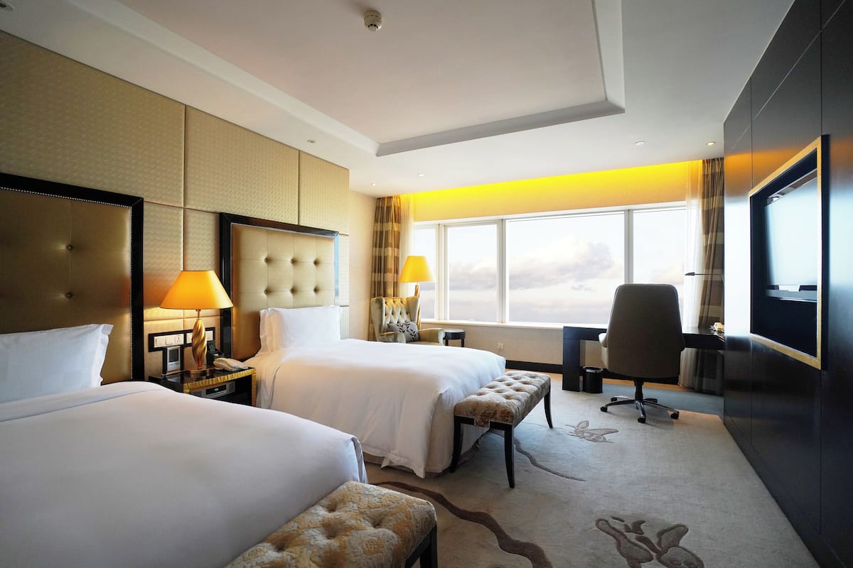 A hotel room with two beds, a desk, a chair, a TV, and a large window.