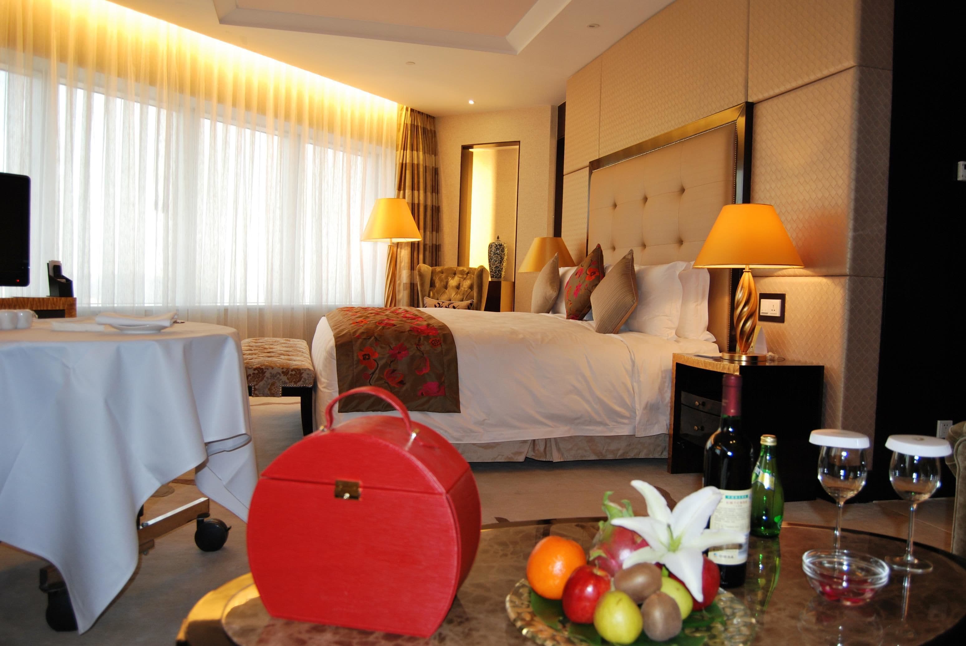 Una habitación de hotel con una cama, lámparas de noche, una mesa con una cesta de frutas, un bolso rojo y un televisor sobre un mueble.