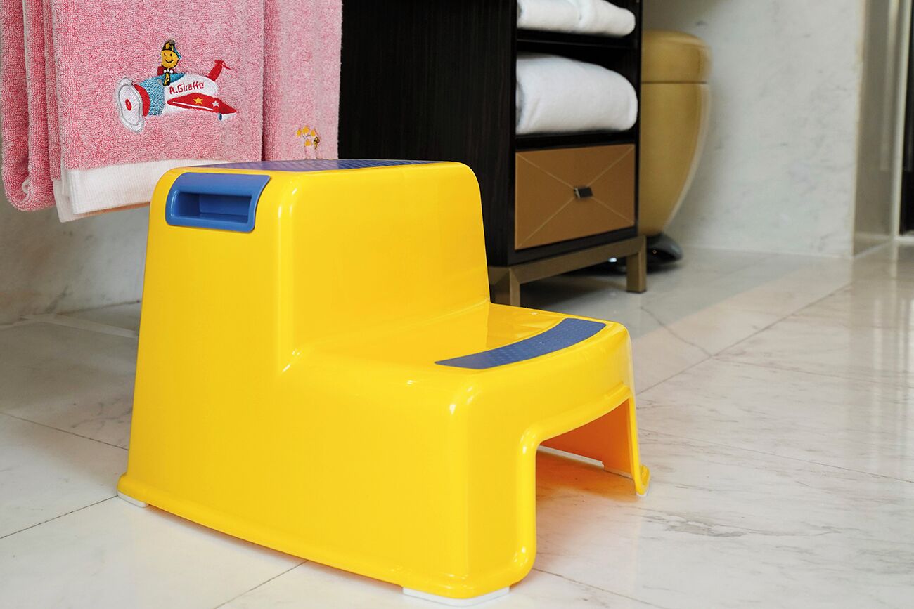 A yellow plastic step stool with a blue top in a room with a tiled floor, a pink towel with a cartoon character, and a dark wooden cabinet with drawers.