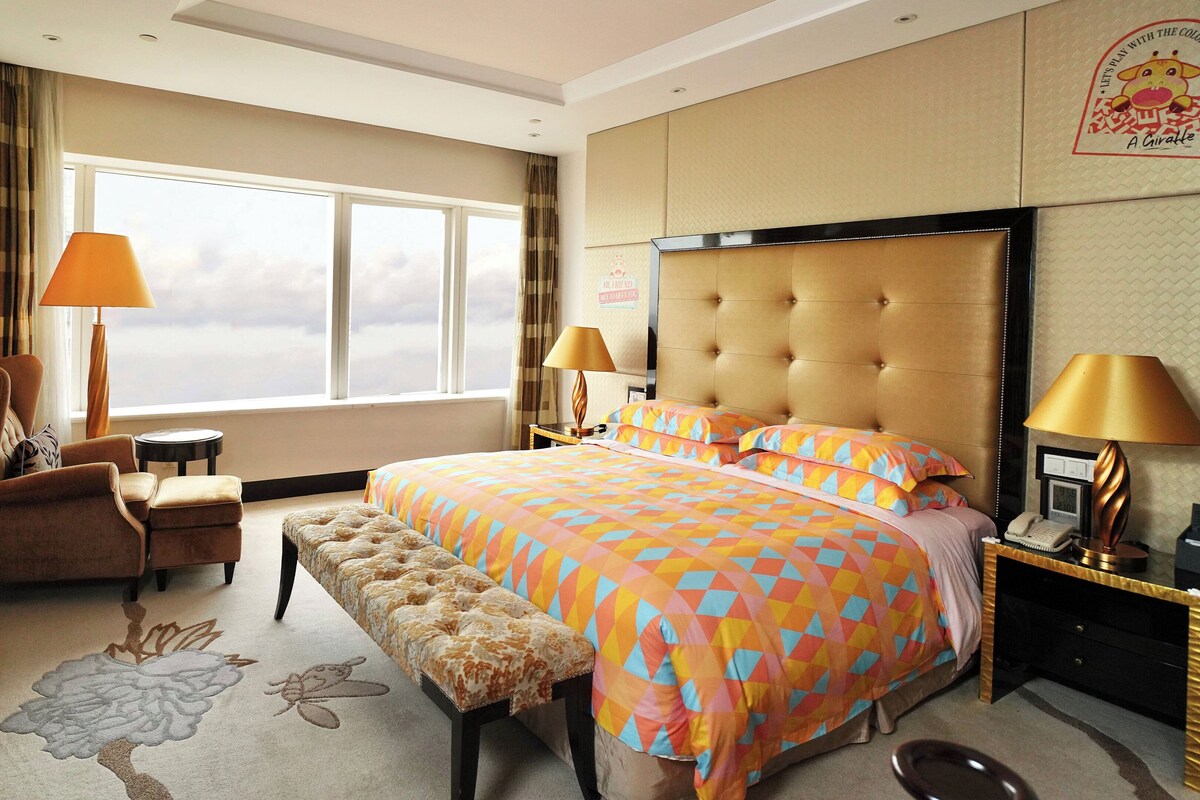 A hotel room with a large bed, a sitting area with a chair and bench, a television, and a window with curtains.