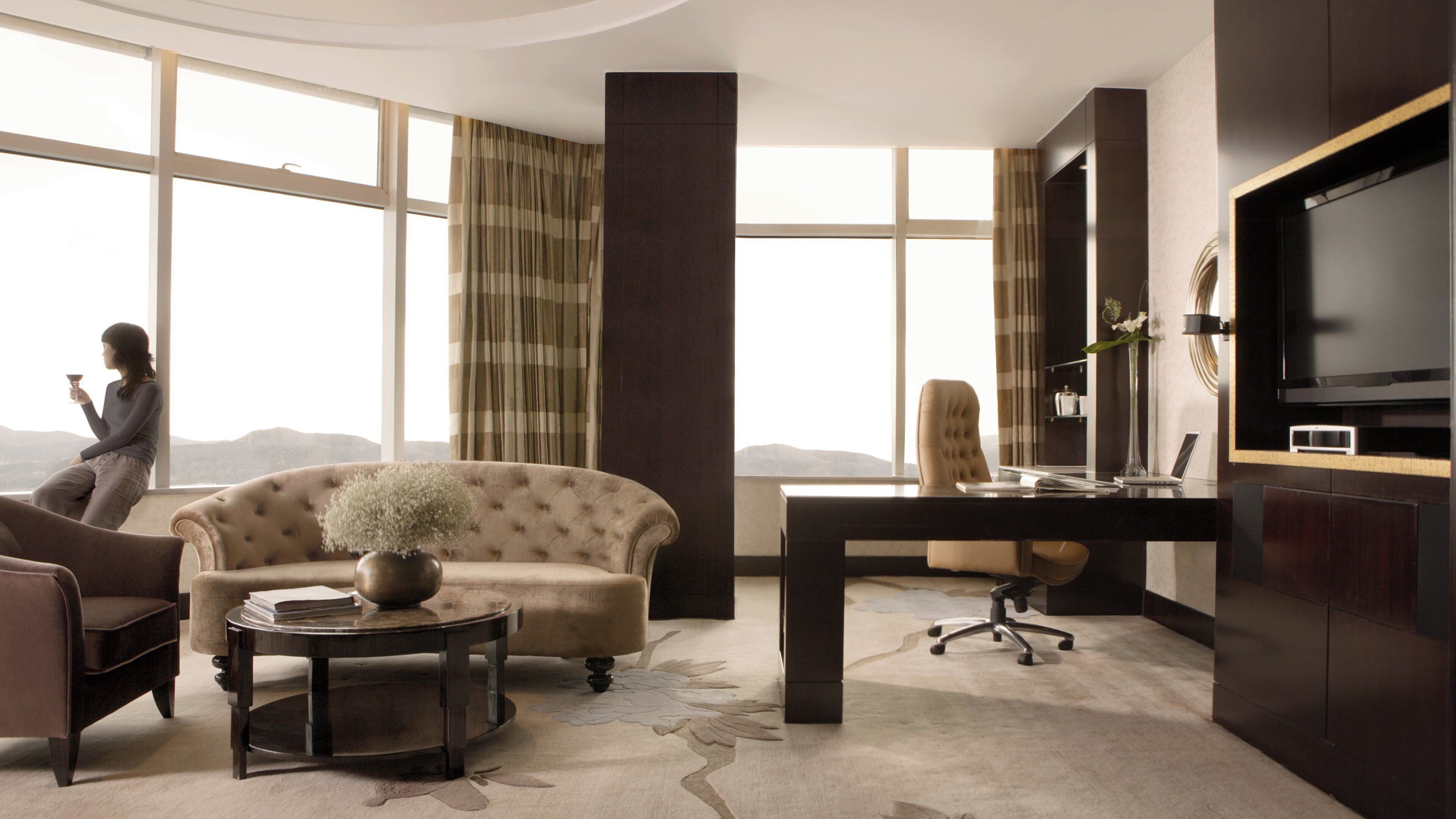 A modern living room with a sofa, a coffee table, a desk, and a television. There is a view of the outdoors through large windows.