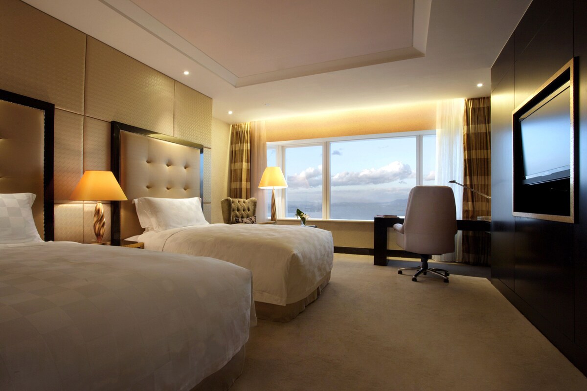 A hotel room with two beds, a desk, a chair, a TV, and a large window with a view of the sky.