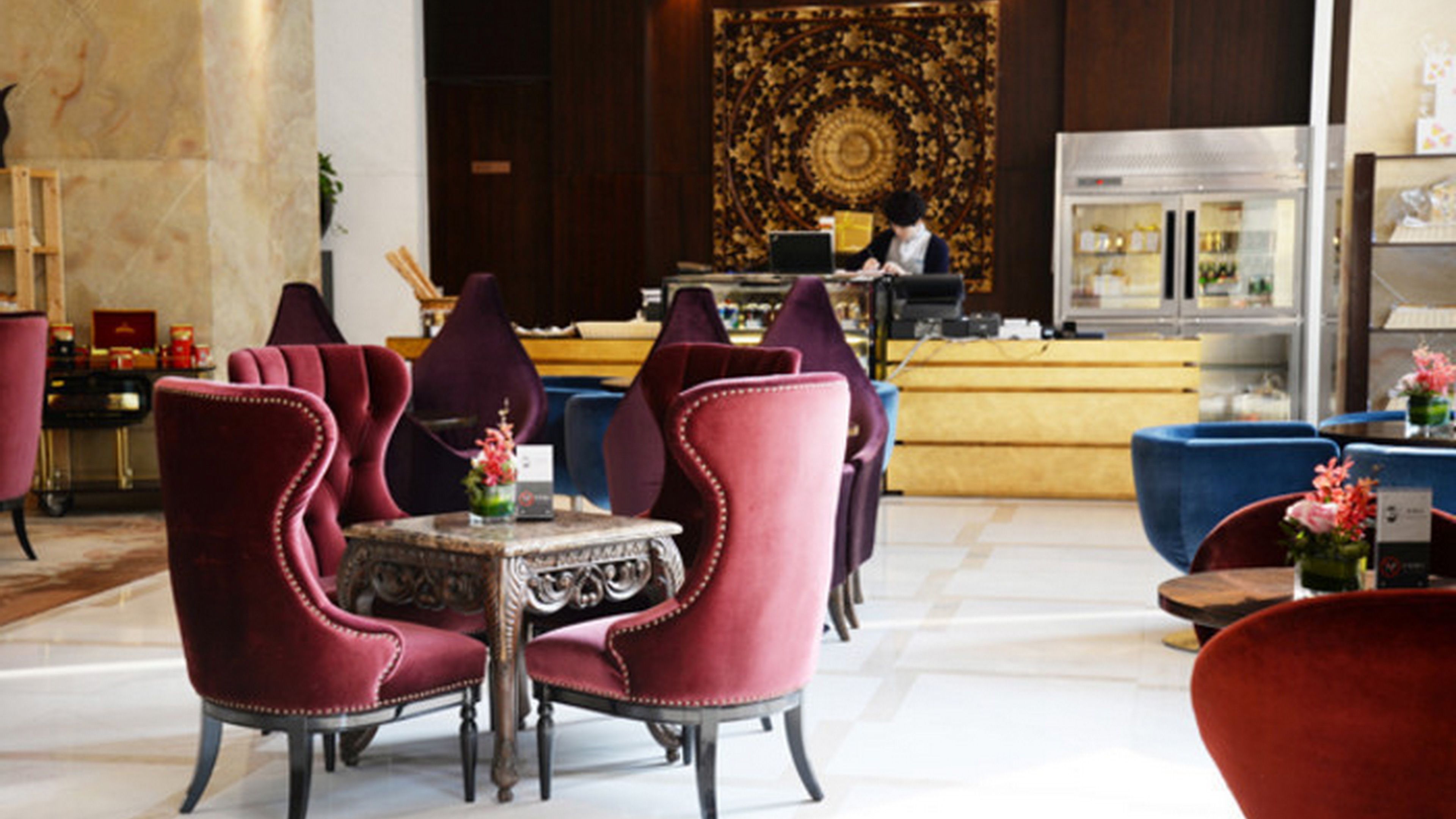 A hotel lobby with red upholstered chairs, a marble floor, a reception desk, and a large decorative tapestry.