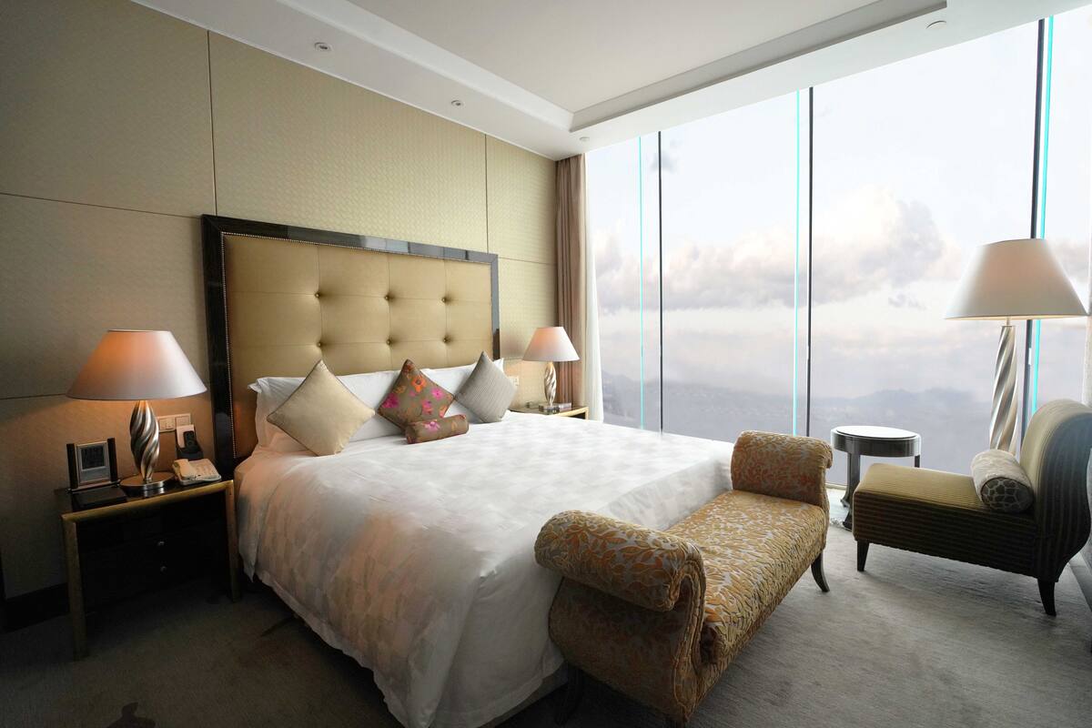 A hotel room with a large bed, two bedside tables, a chair, a small round table, and a view of the sky through large windows.
