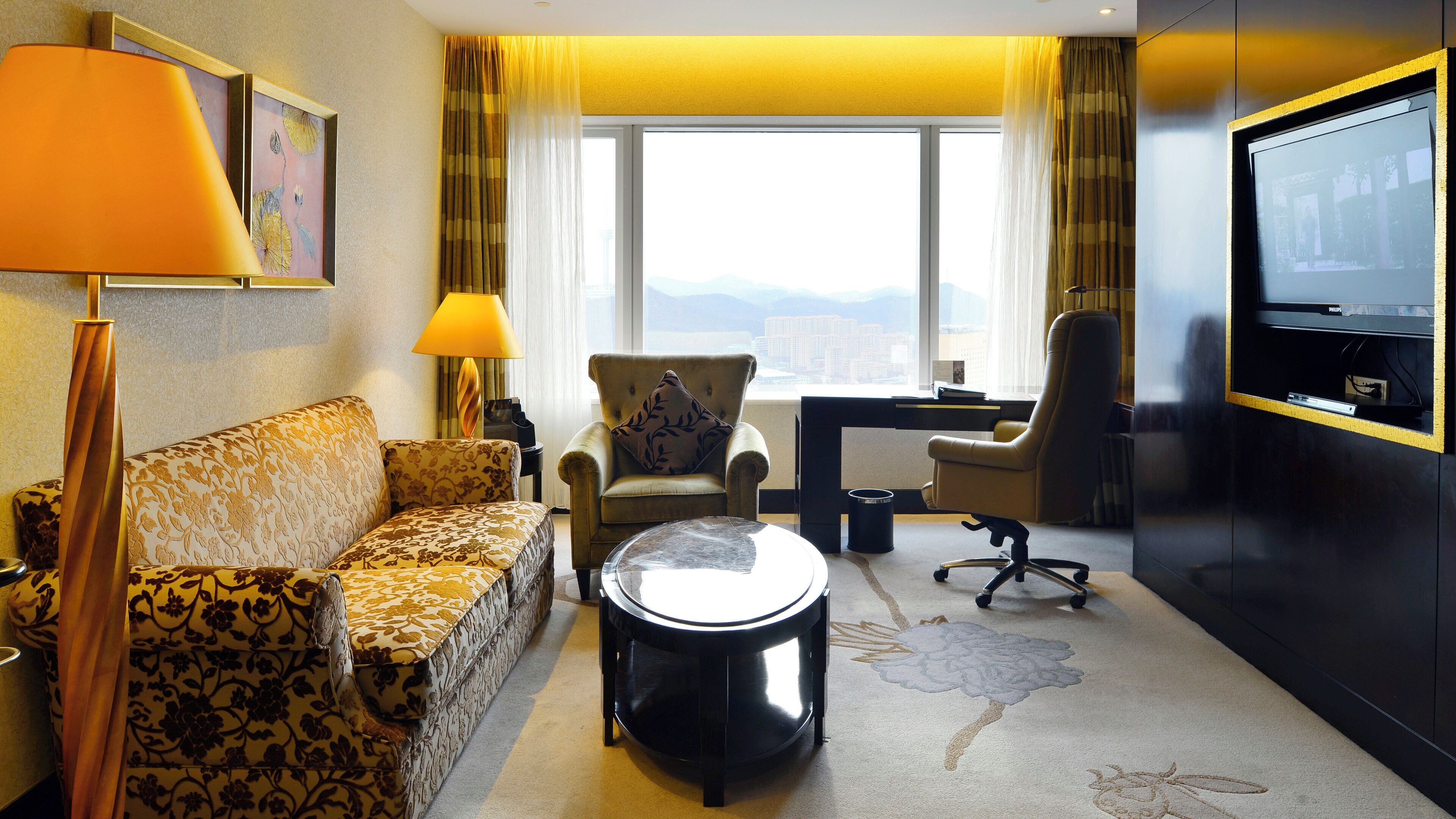 Habitación de hotel con sofá, sillón, escritorio, silla, televisor y vistas a la ciudad.