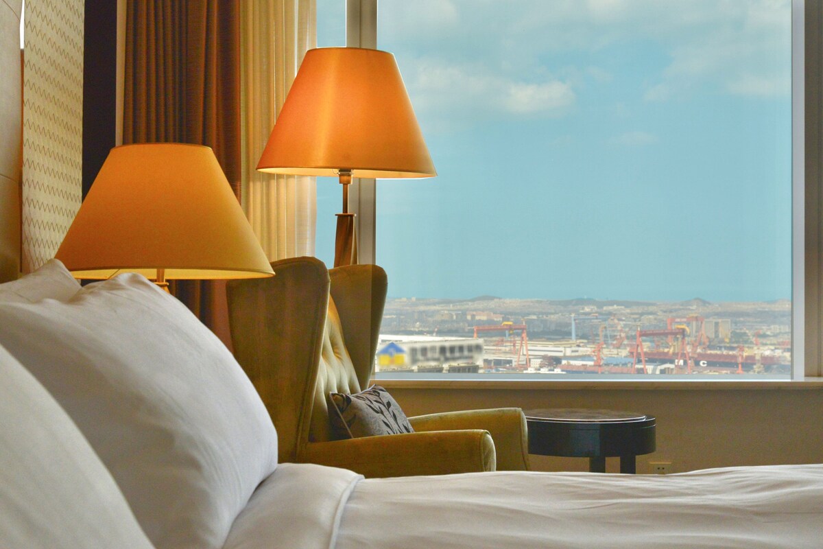 Ein Hotelzimmer mit einem Bett, zwei Lampen, einem Sessel und Blick auf eine Stadtlandschaft durch ein Fenster.