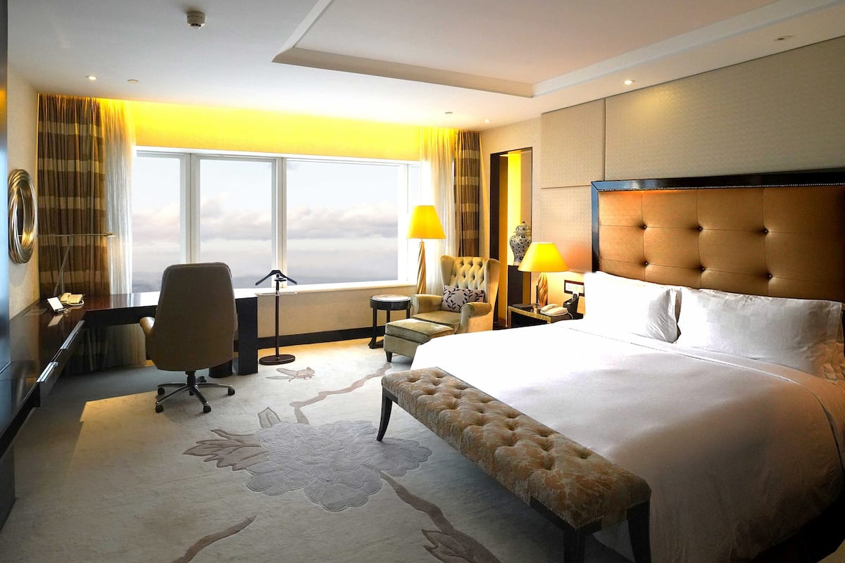A hotel room with a large bed, a desk with a chair, a TV, and a window with a view of the sky.