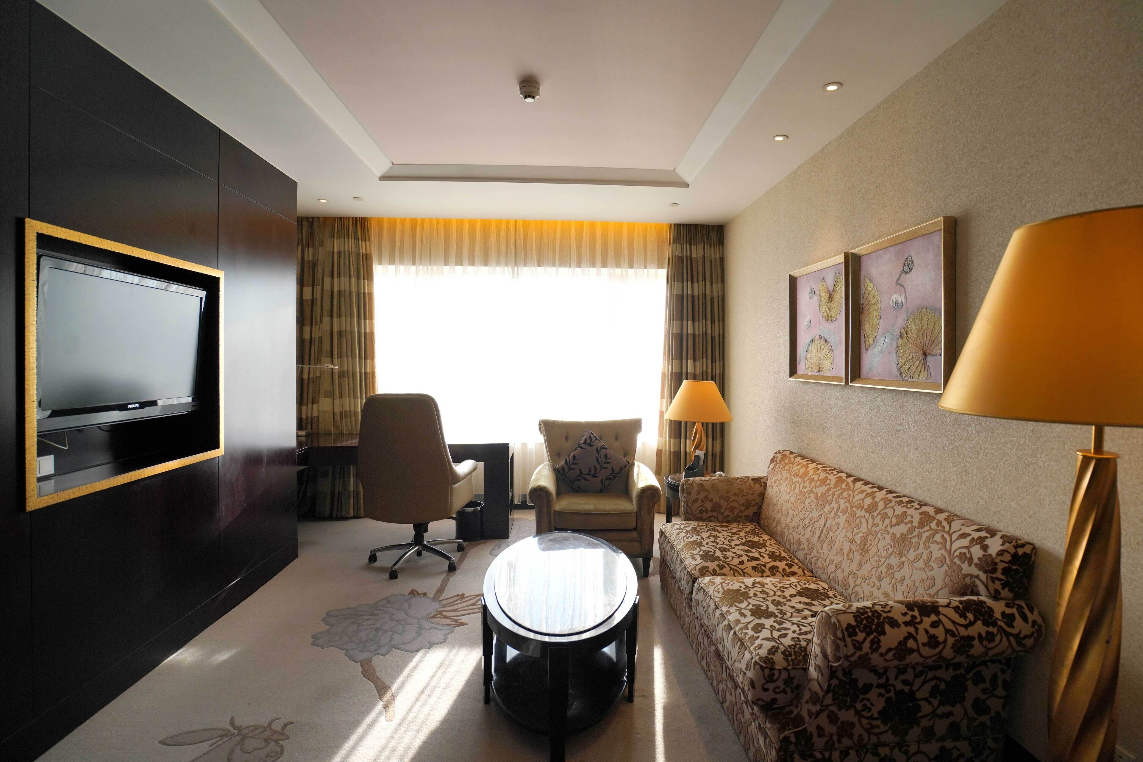 A hotel room with a flat-screen TV, a sofa, a chair, a coffee table, and a lamp.