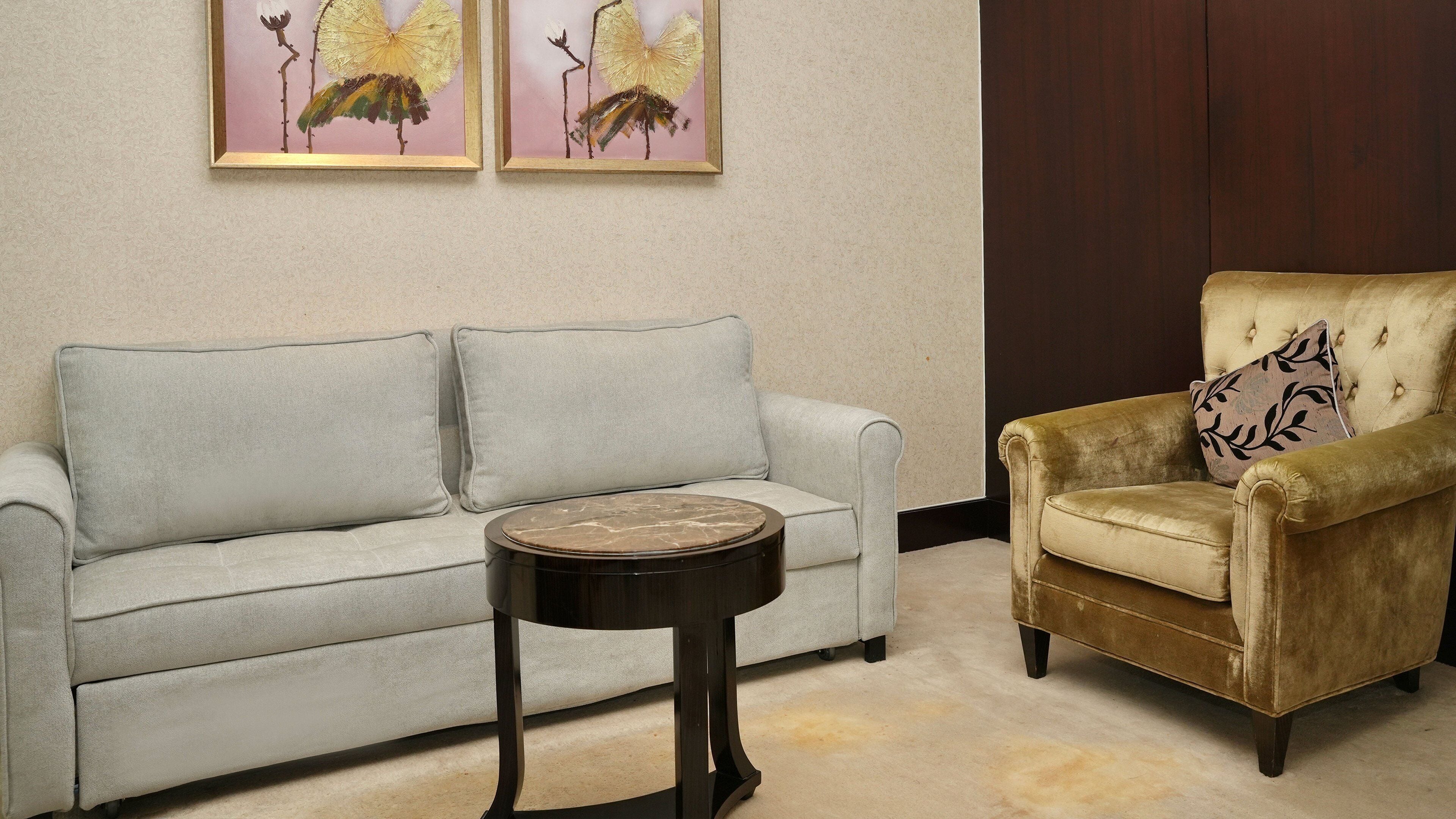 A living room with a light grey sofa, a round side table, and a gold armchair with a patterned pillow. Two framed artworks are hung on the wall.