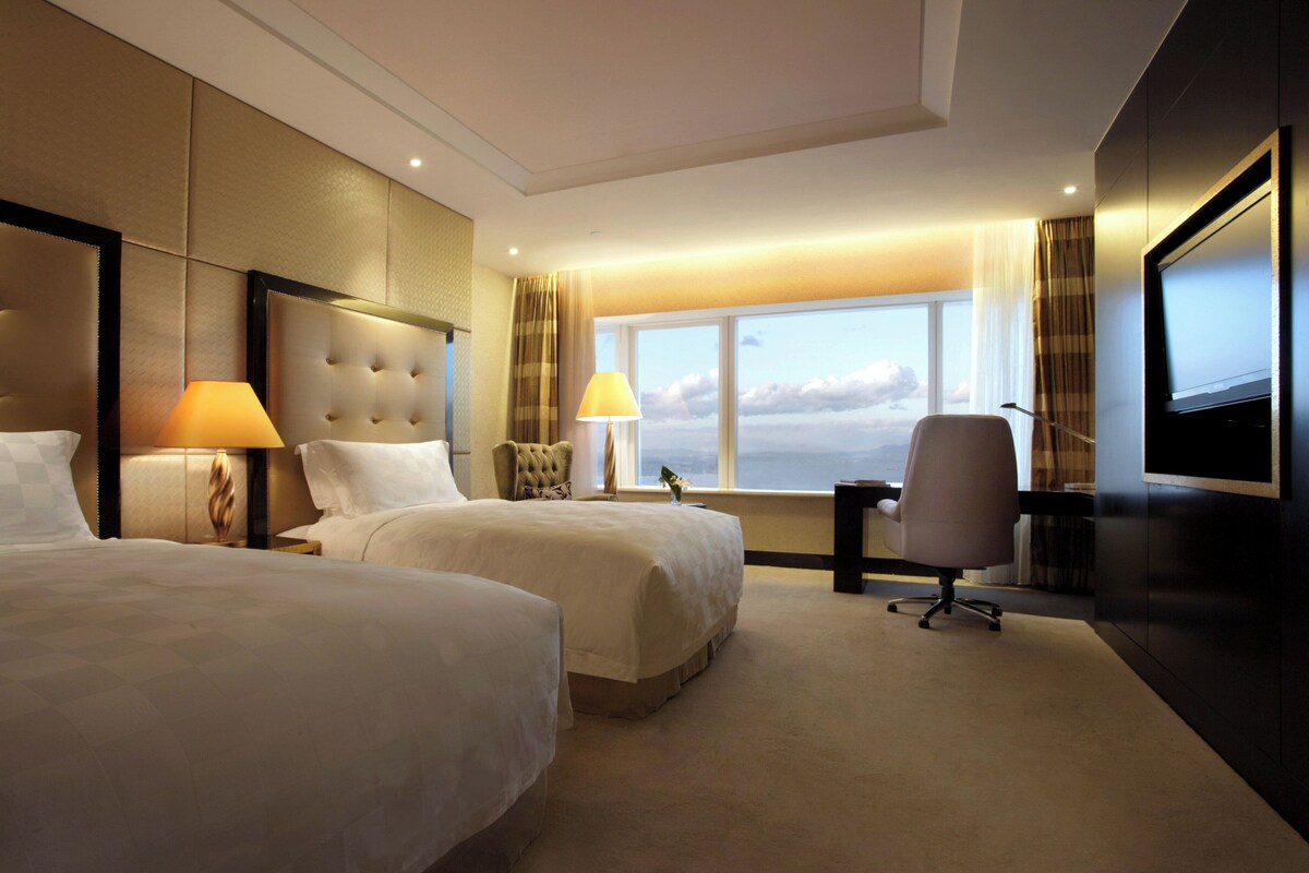 A hotel room with two beds, a desk, a chair, a television, and a large window with a view.