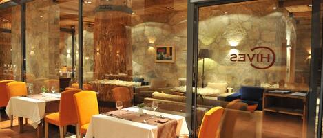 A restaurant with wooden interior, stone walls, and orange chairs. Tables are set with white tablecloths and wine glasses.