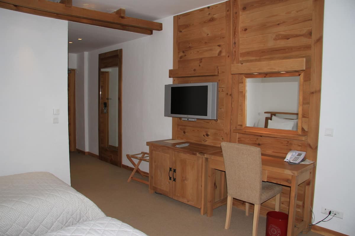 Quarto com cama, escrivaninha de madeira, cadeira, televisão e espelho.