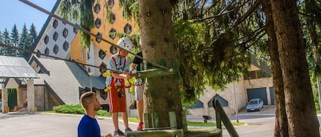 Una persona cruzando un puente colgante en un parque, con árboles y un edificio al fondo.