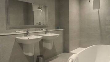 A modern bathroom with two sinks, a large mirror, a bathtub, and a trash can.