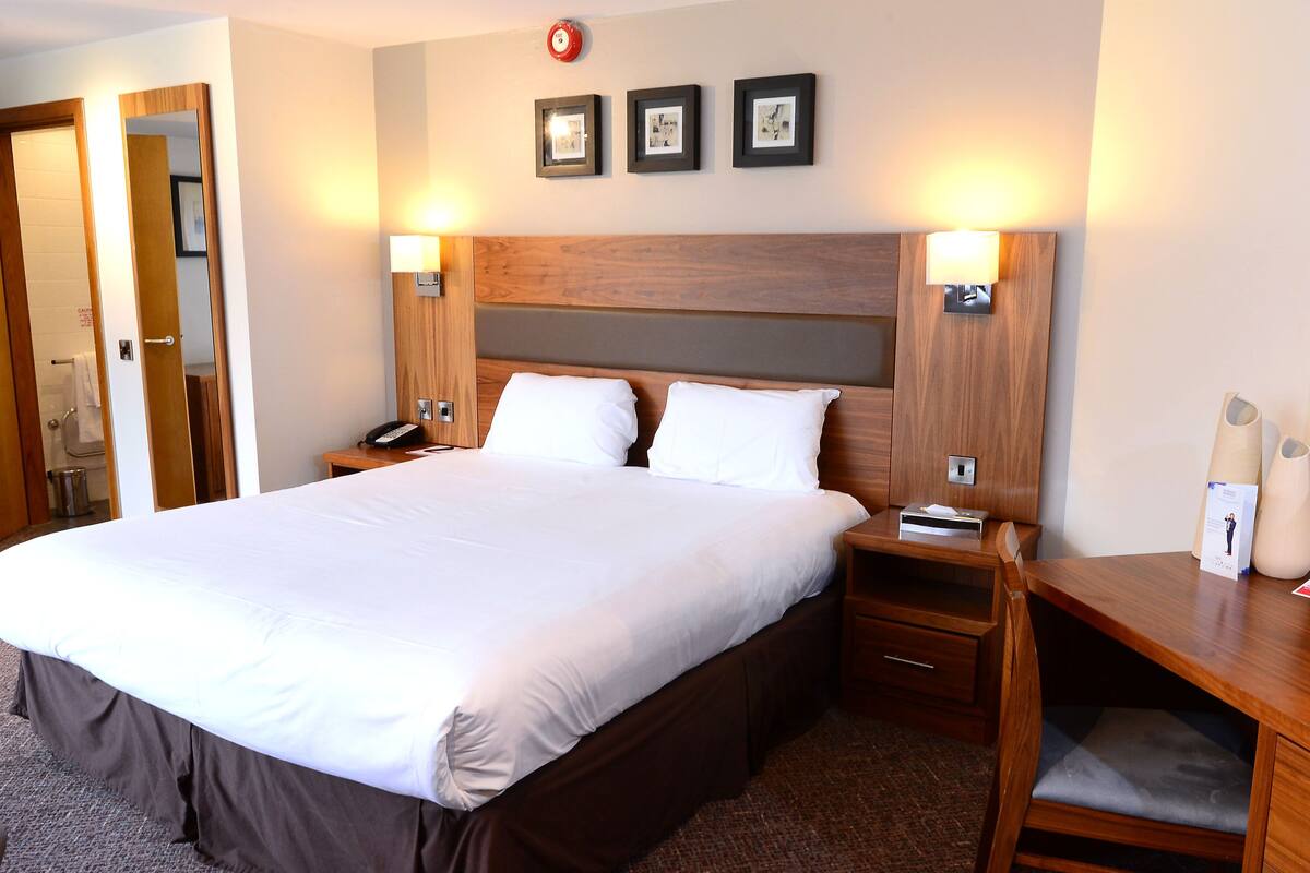 A hotel room with a large bed, wooden headboard, bedside tables, and a nightstand with a lamp.