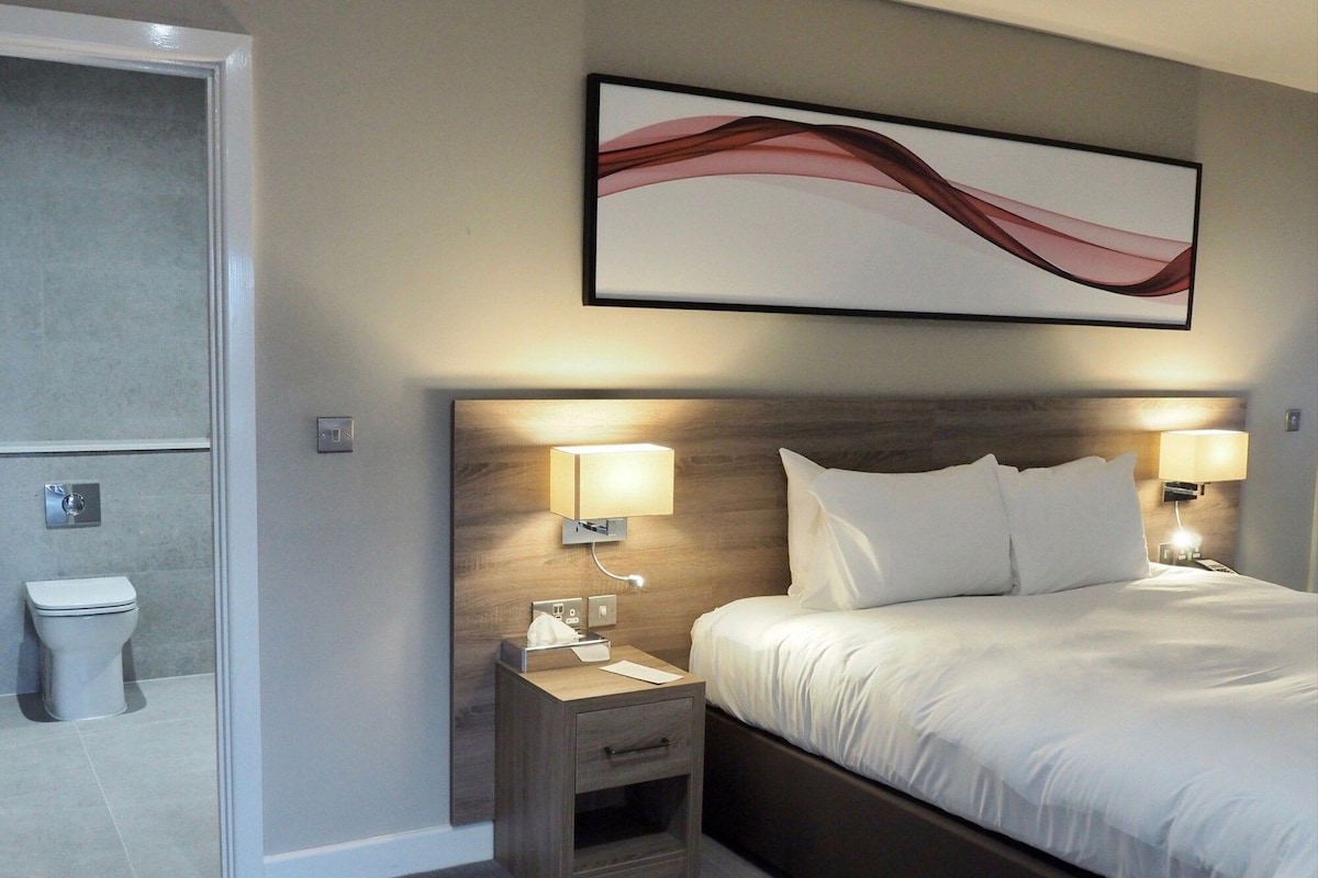 A hotel room with a bed, bedside lamps, a nightstand, a wooden headboard, a wall-mounted abstract art piece, and a toilet visible through an open door.