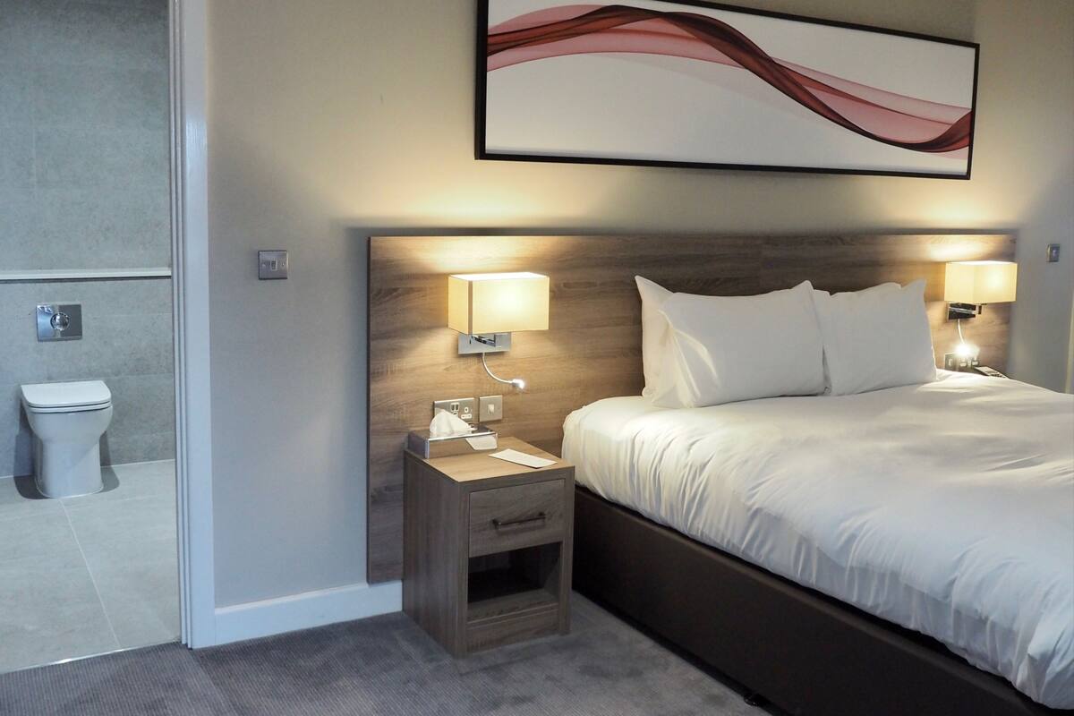 Une chambre d’hôtel avec un lit, des lampes de chevet, une table de nuit, une tête de lit en bois, une œuvre d’art abstraite accrochée au mur et des toilettes visibles à travers une porte ouverte.