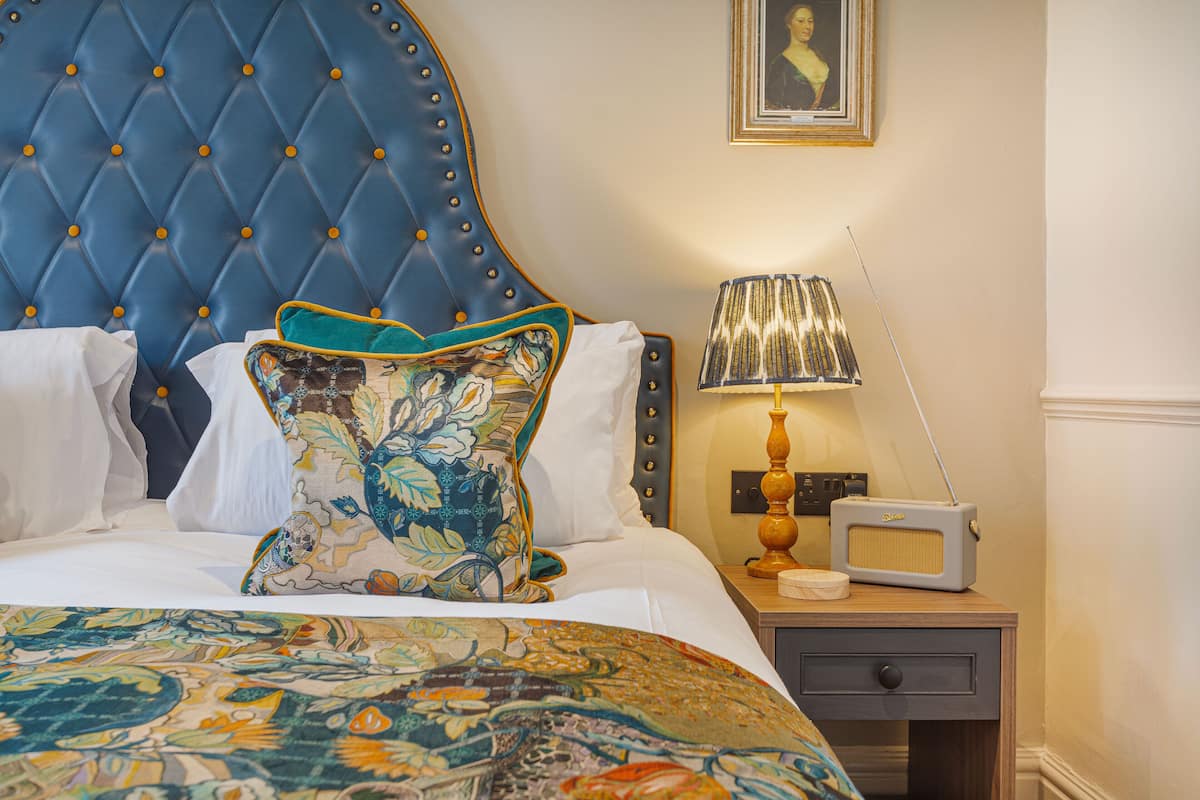 A hotel room with a tufted headboard, a patterned throw blanket, a bedside lamp, and a vintage radio.