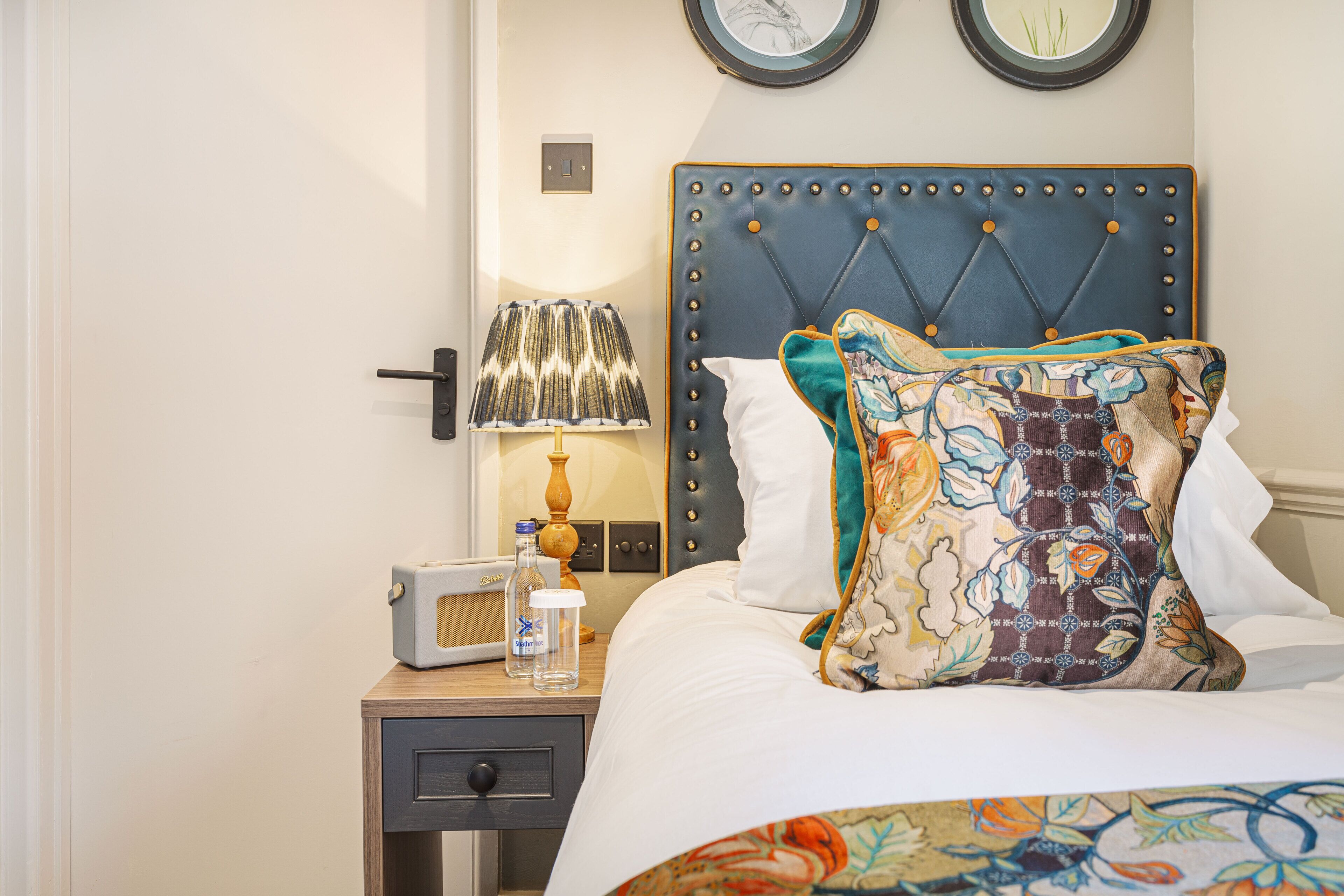 A neatly made bed with a decorative pillow, a bedside lamp, a radio, and a small table with a bottle and glass.