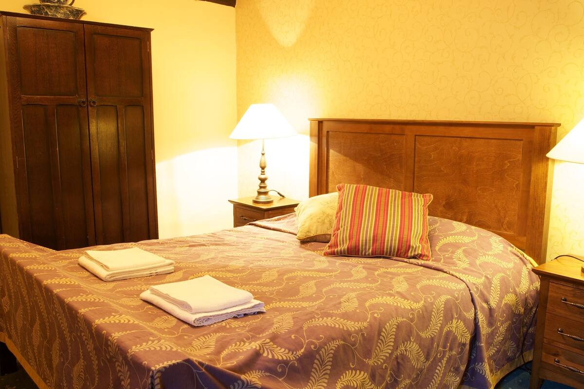 A hotel room with a bed, a wooden wardrobe, a nightstand with a lamp, and a bedside table.