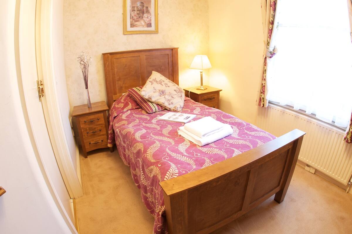 A bedroom with a wooden bed, a nightstand with a lamp, a vase with dried flowers, and a radiator.