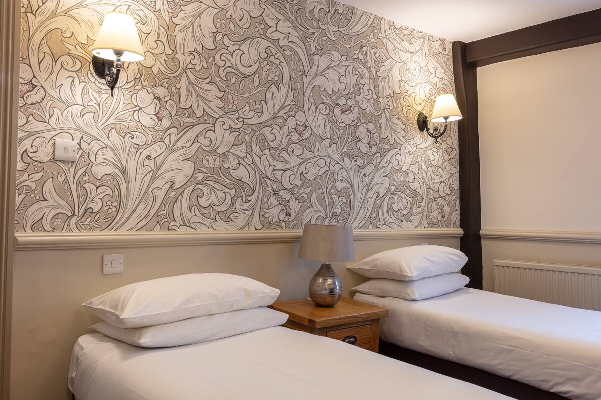 A hotel room with two beds, a patterned wallpaper, and wall-mounted lamps.