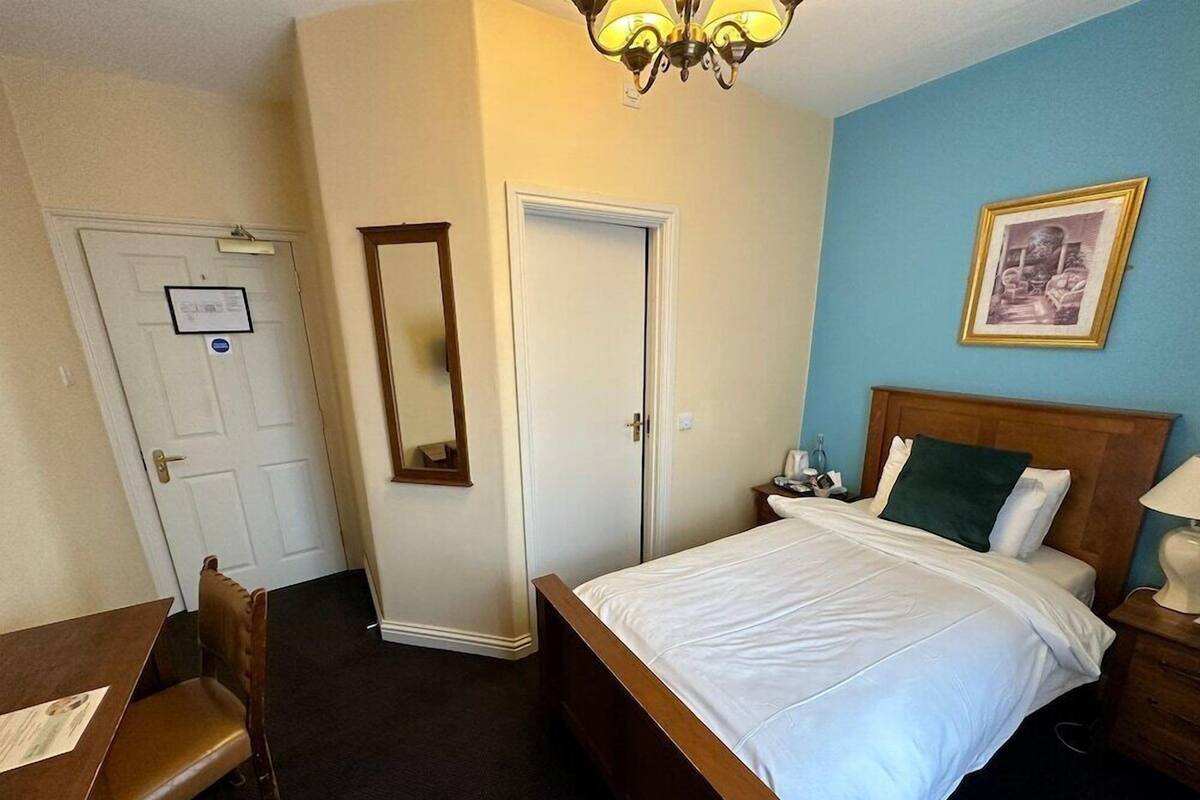 A hotel room with a bed, a desk, a chair, a mirror, a door, and a framed picture on the wall.