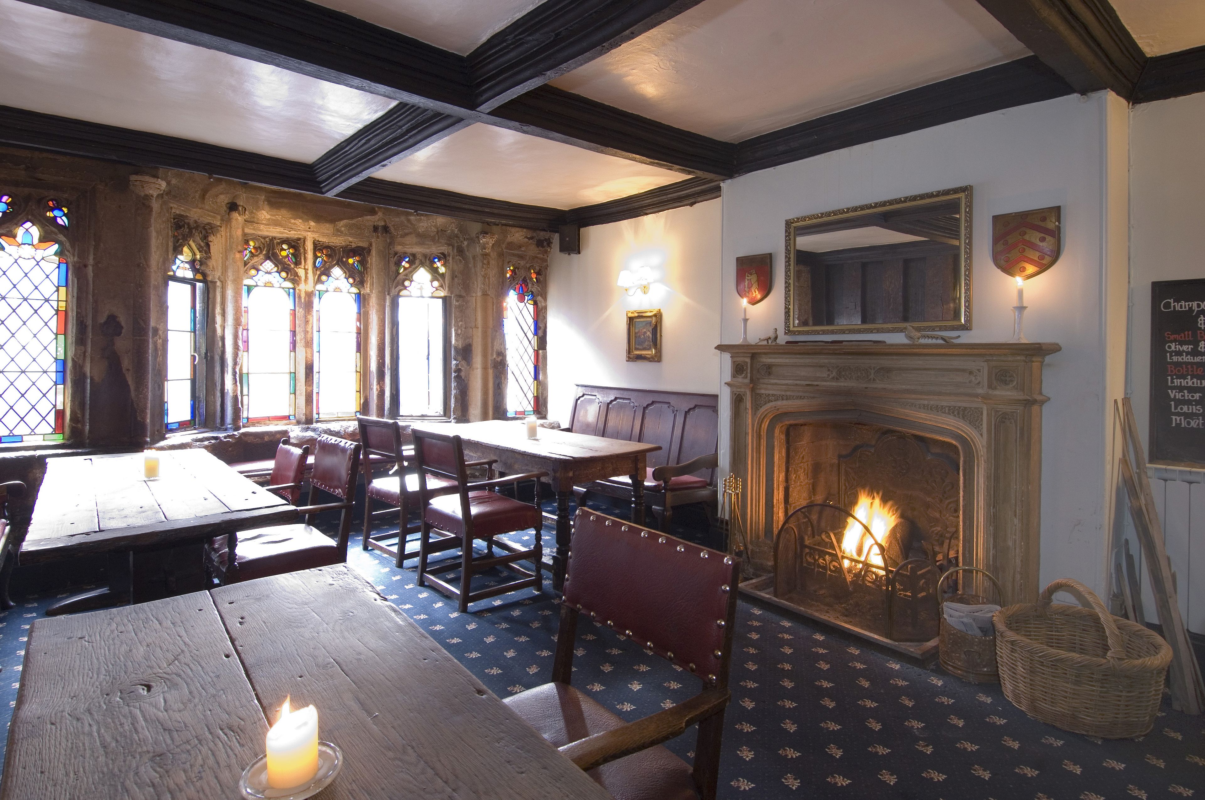 A traditional inn room with wooden furniture, a fireplace, stained glass windows, and a stone wall.