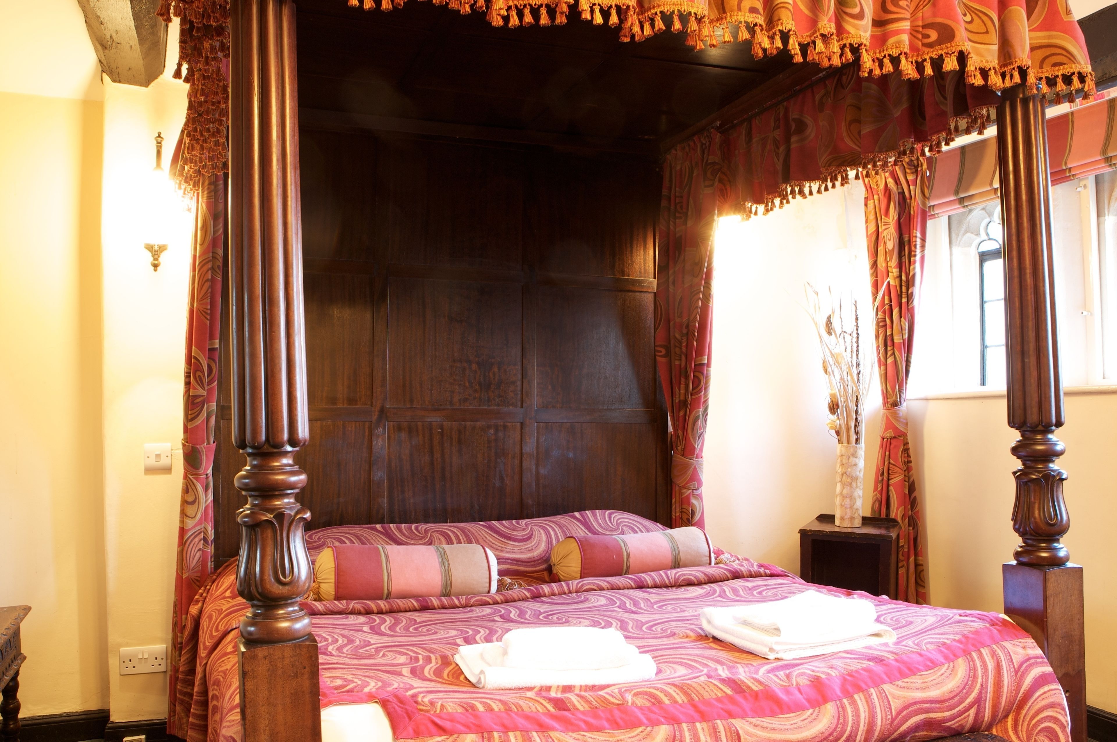 A four-poster canopy bed with a wooden headboard, a patterned bedspread, and a bedside table with a vase.
