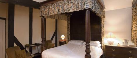 A four-poster bed with a floral canopy, a wooden nightstand with a lamp, a wooden dresser with drawers, and a chair with a cushion.