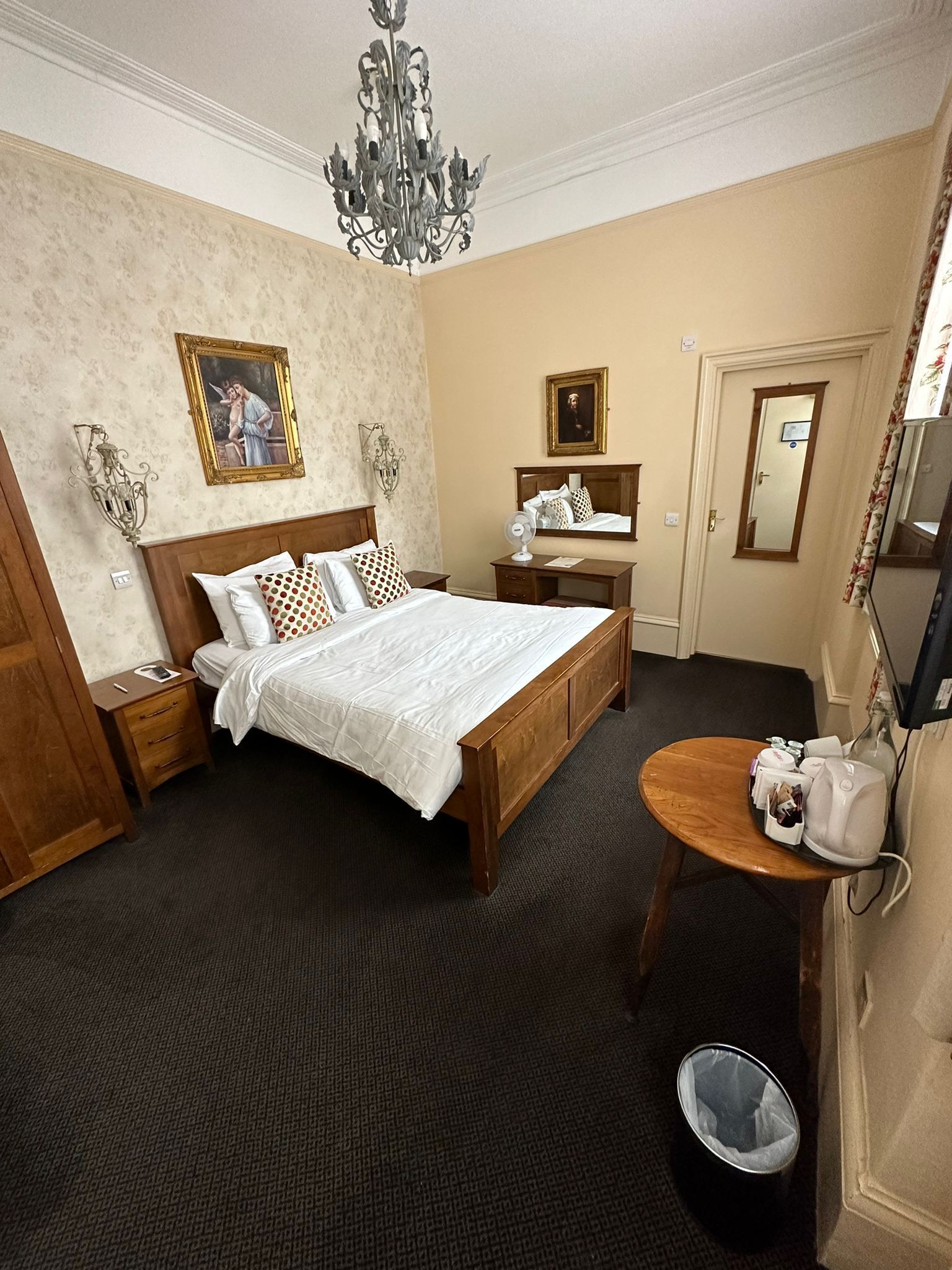 Une chambre d’hôtel avec un lit en bois, une table de chevet, une petite table ronde avec une bouilloire, un miroir et un lustre.