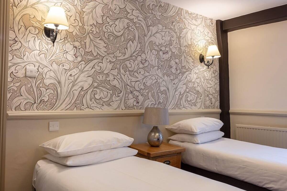 A hotel room with two beds, a patterned wallpaper, wall-mounted lamps, and a nightstand with a lamp.