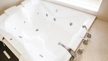 Una bañera blanca grande con múltiples grifos y un desagüe, ubicada en un baño con piso de baldosas y una ventana con persianas.