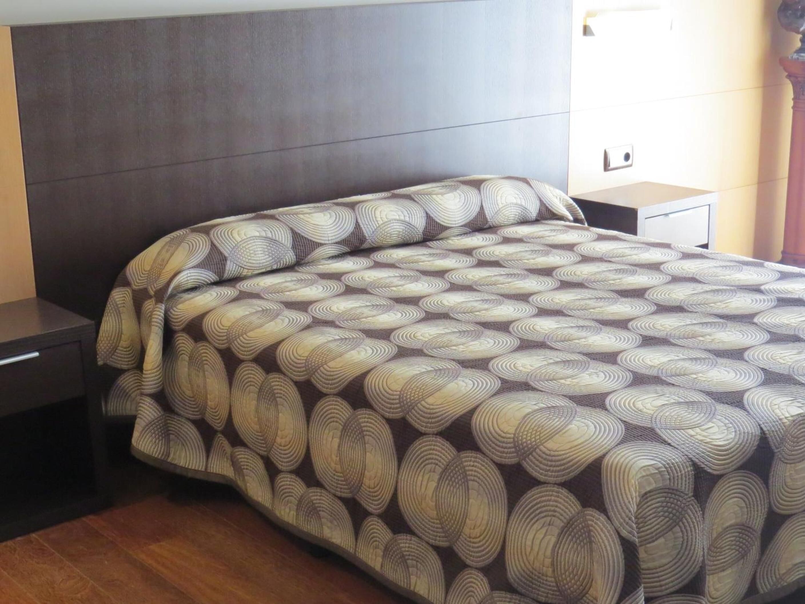 A bedroom with a dark headboard, a patterned bedspread, a nightstand, and a bedside table.