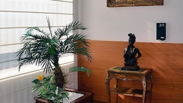 Een kamer met een groot raam, een potplant, een lijst met een schilderij en een tafeltje met een beeldhouwwerk.