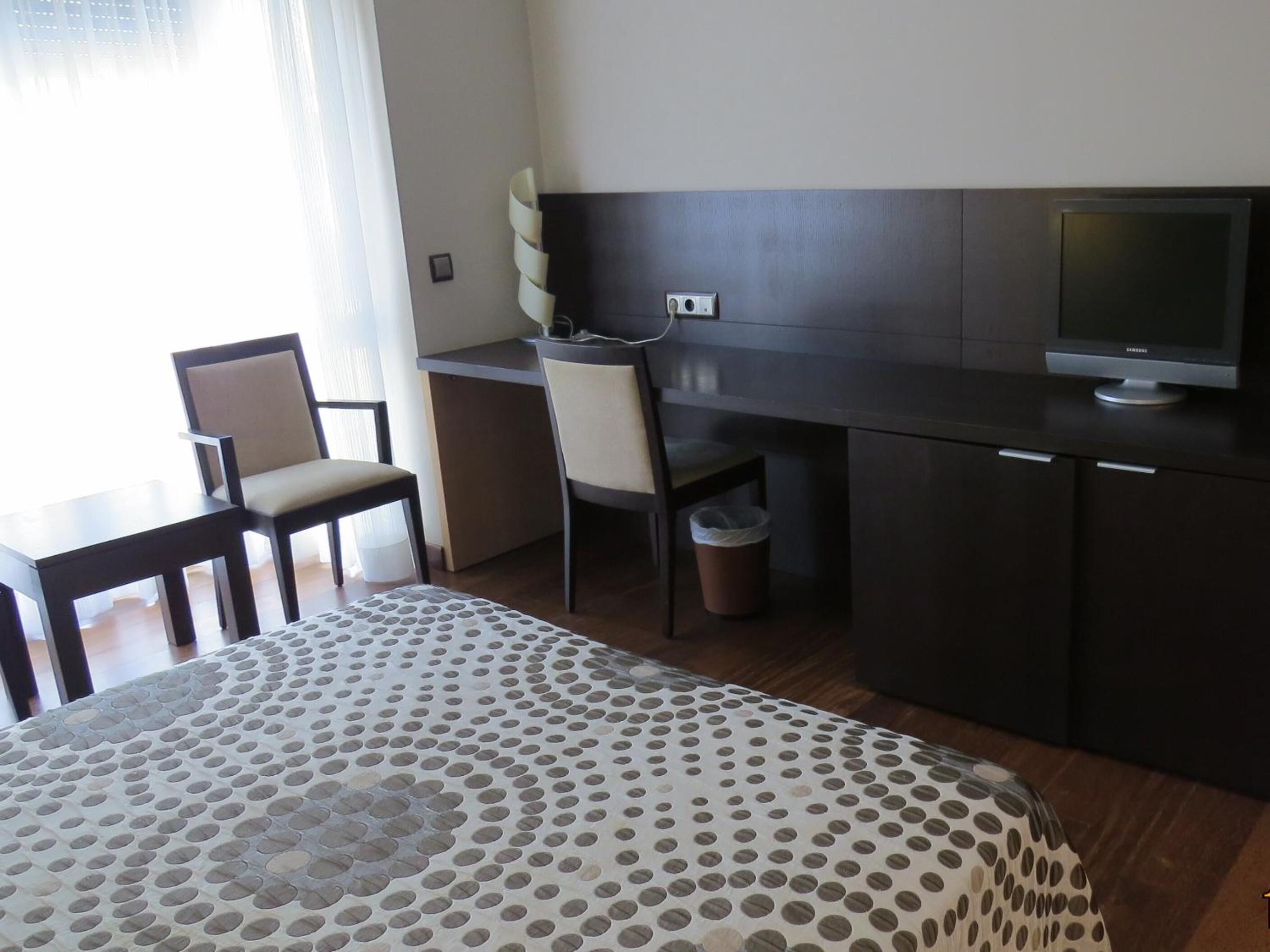 A hotel room with a bed, a desk, two chairs, a television, and a waste bin.