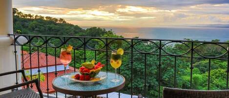 A balcony with a table set for two, overlooking a lush landscape and the sea.