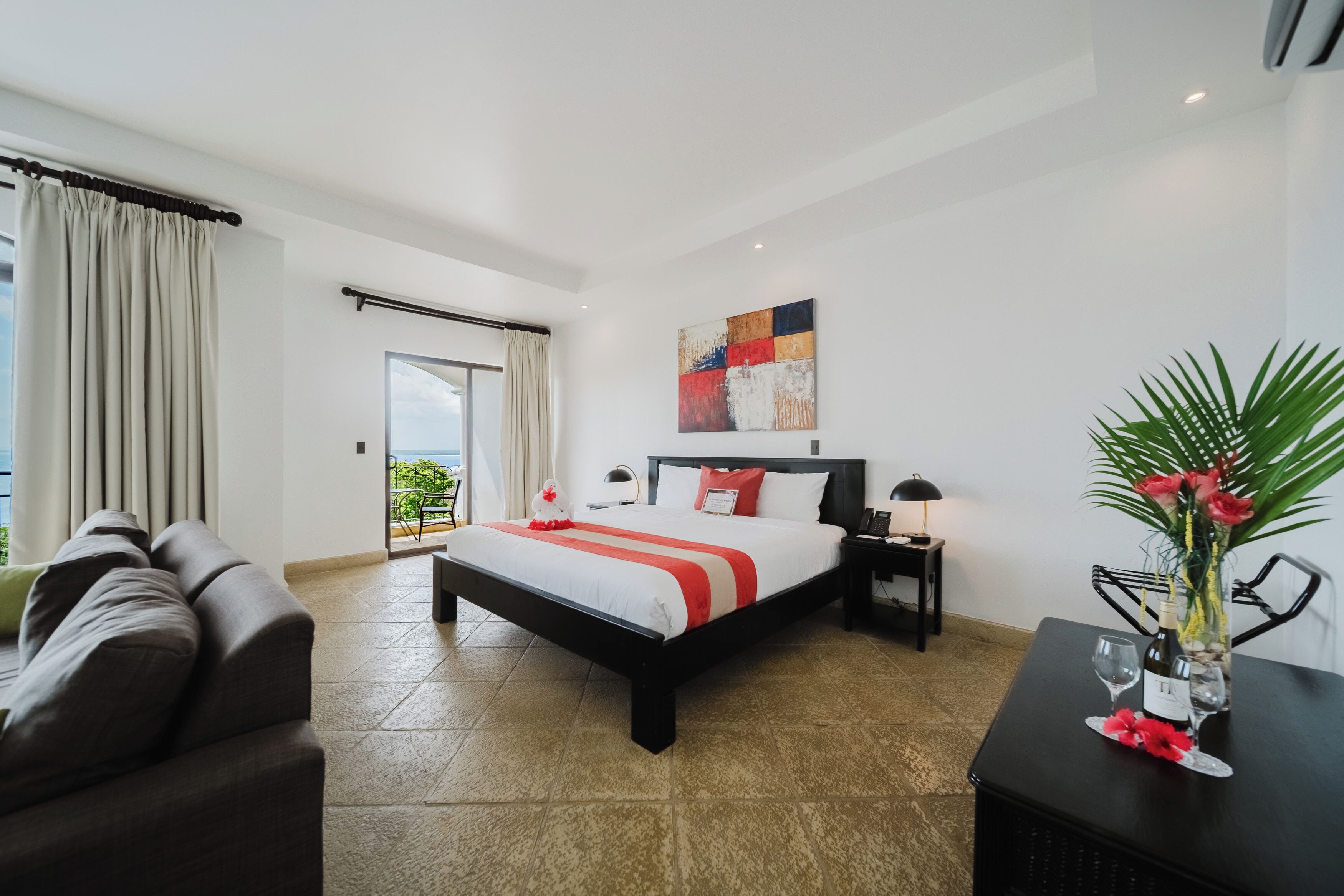 A hotel room with a bed, a sofa, a nightstand, a lamp, a vase with flowers, and a view of the outdoors.
