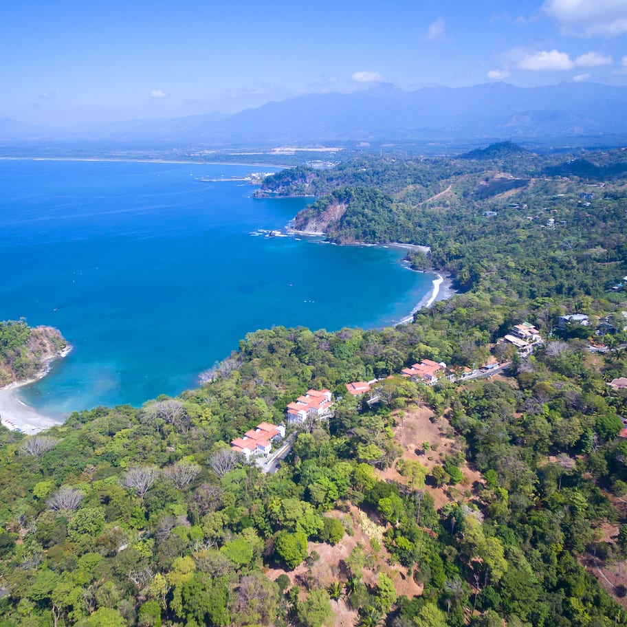 An aerial view of a coastal area with a beach, dense forest, and scattered buildings.
