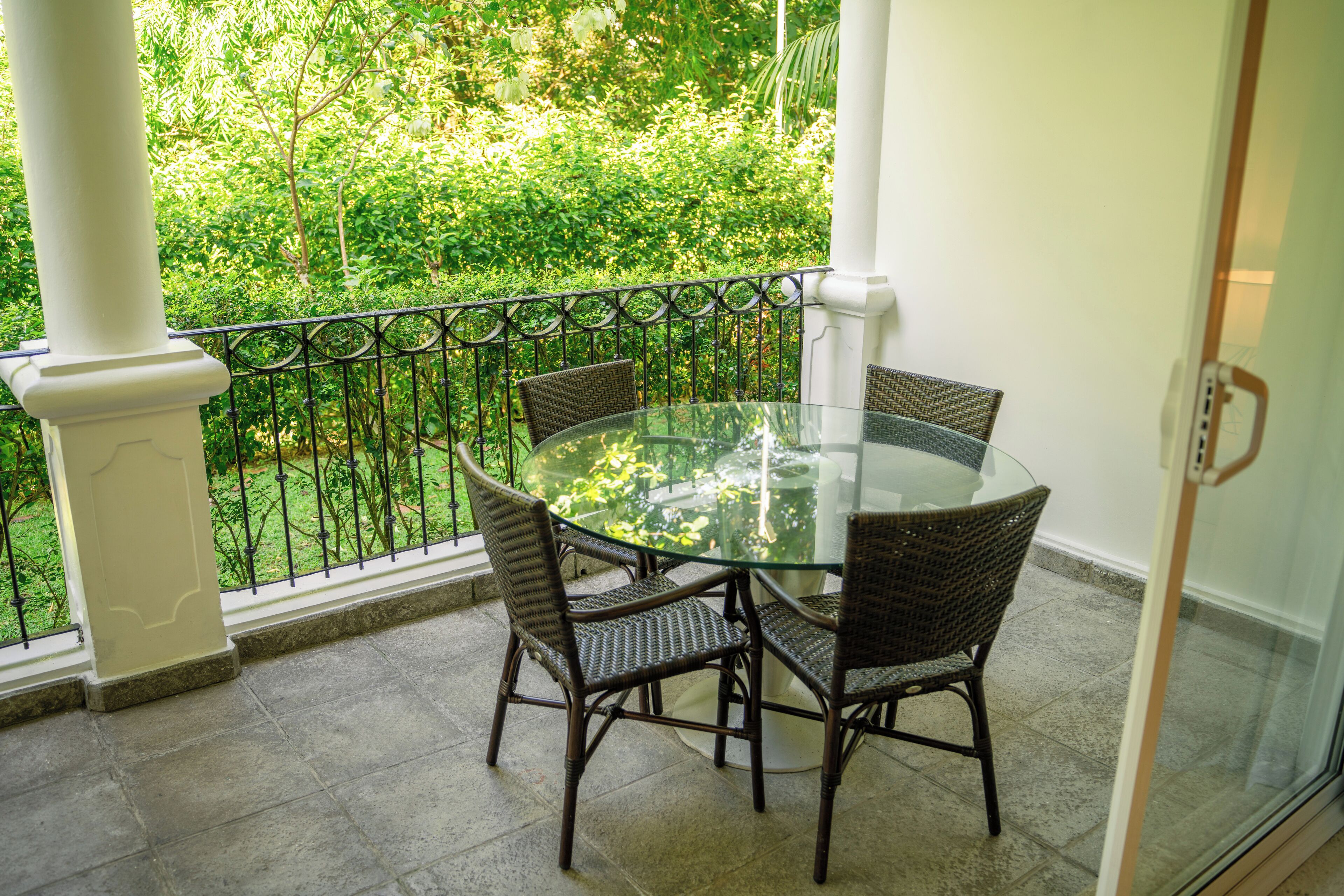 A glass-top table with wicker chairs on a balcony with a view of greenery.