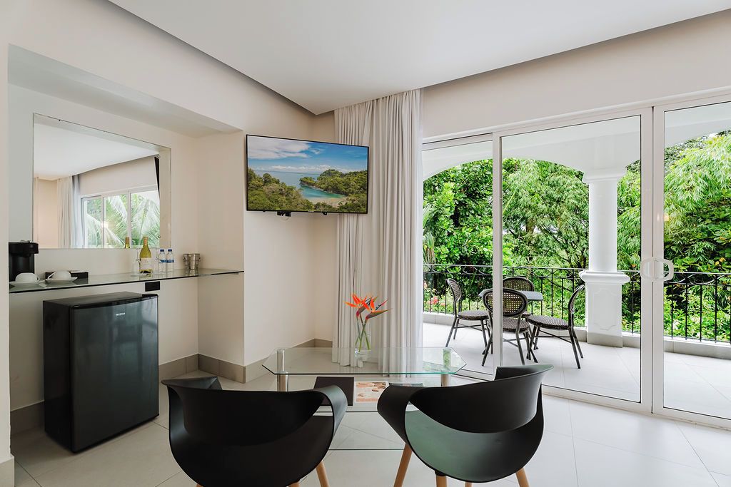 Un salón moderno con una mesa de vidrio, dos sillas negras, un televisor de pantalla plana y un balcón con vistas a zonas verdes.