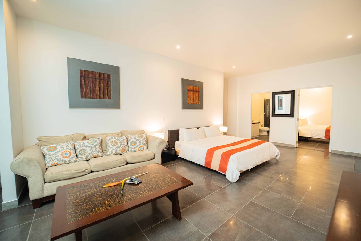 A modern hotel room with a sofa, a bed with an orange bedspread, a coffee table, and two wall-mounted artworks.