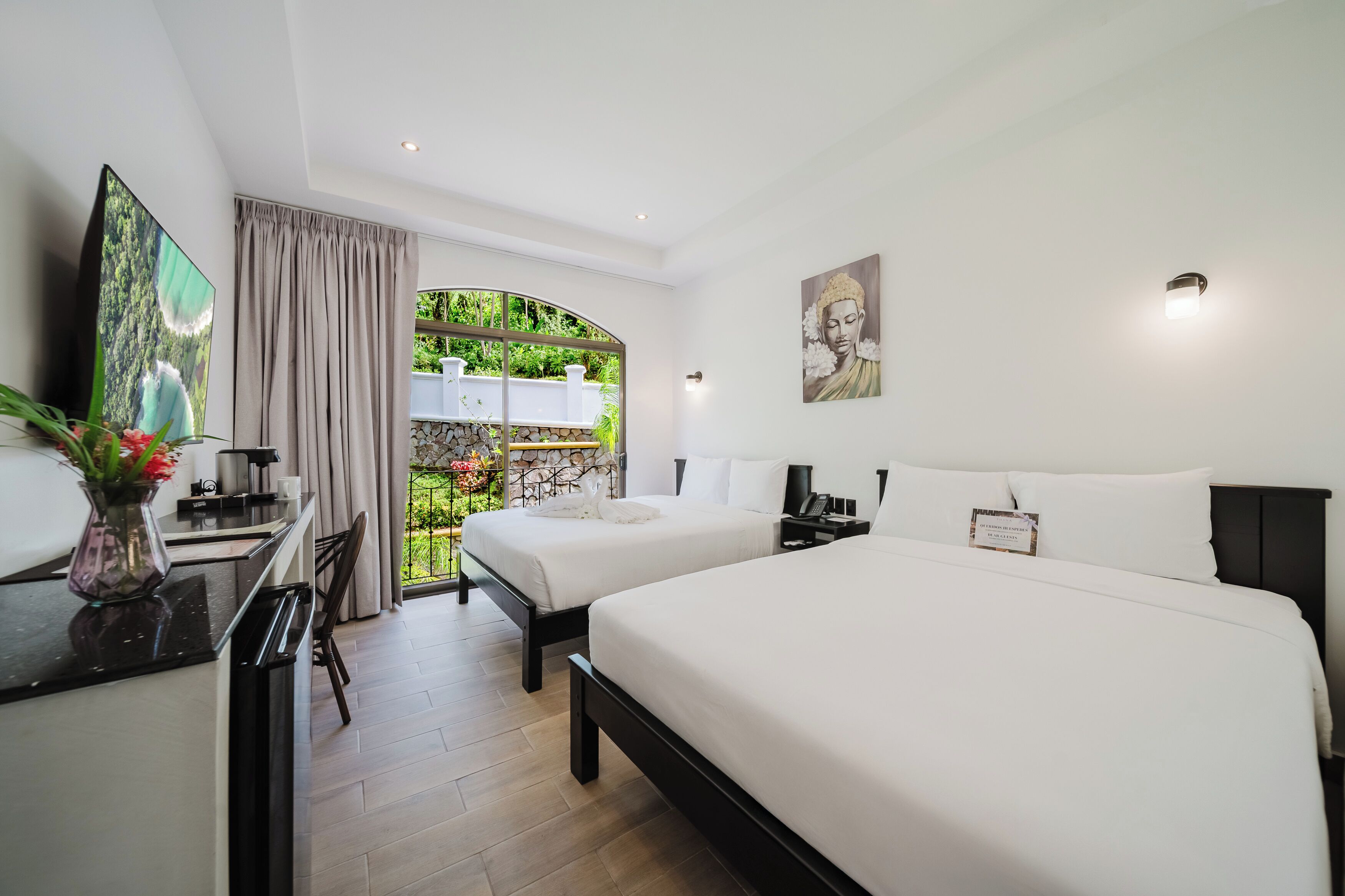 Habitación de hotel con dos camas, un escritorio con televisor, un jarrón con flores y vistas al exterior con vegetación.