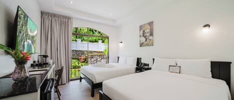 Habitación de hotel con dos camas, un escritorio con televisor, un jarrón con flores y vistas al exterior con vegetación.