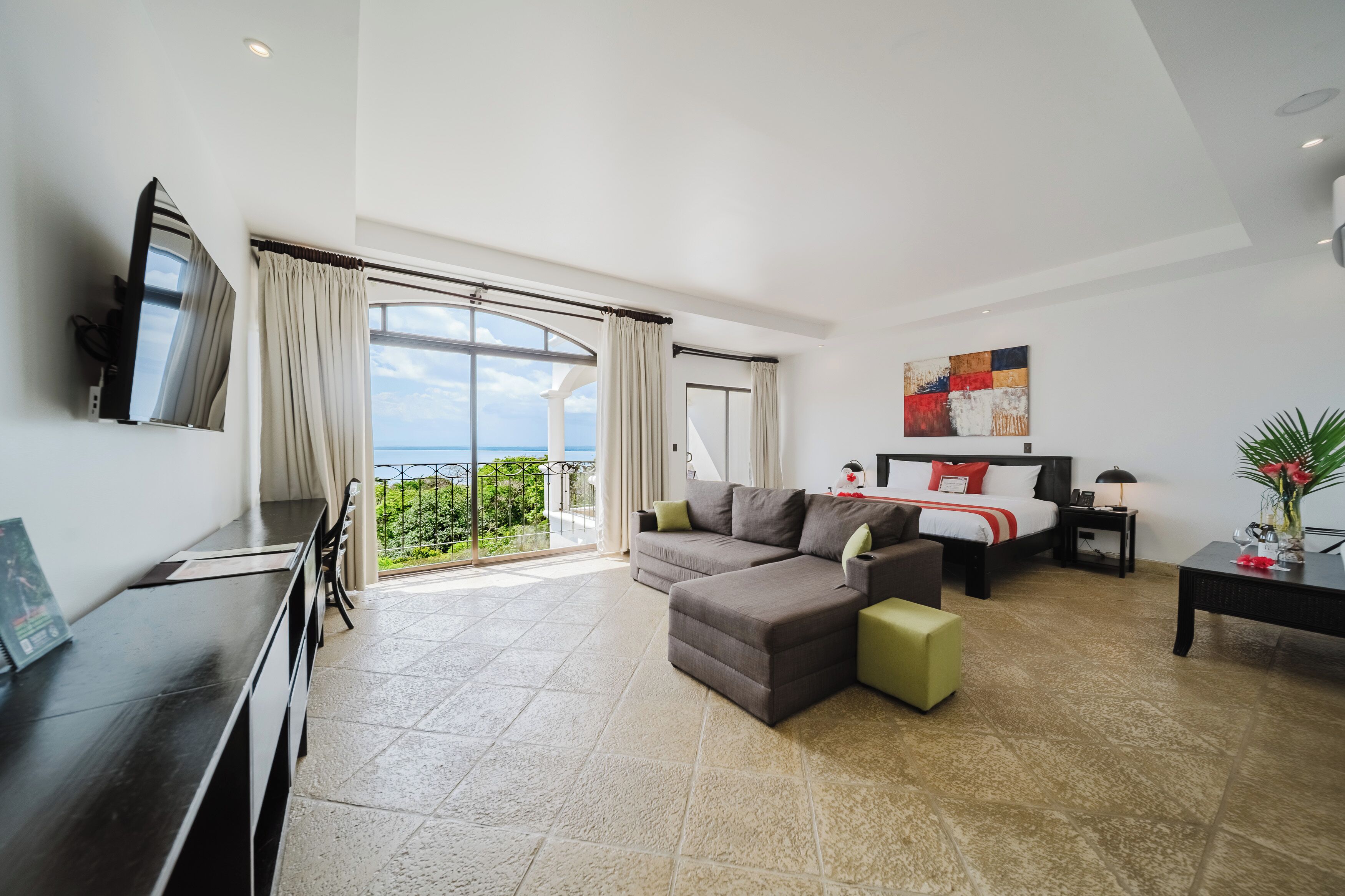 A modern hotel room with a large bed, a sofa, a desk, and a balcony with a view.