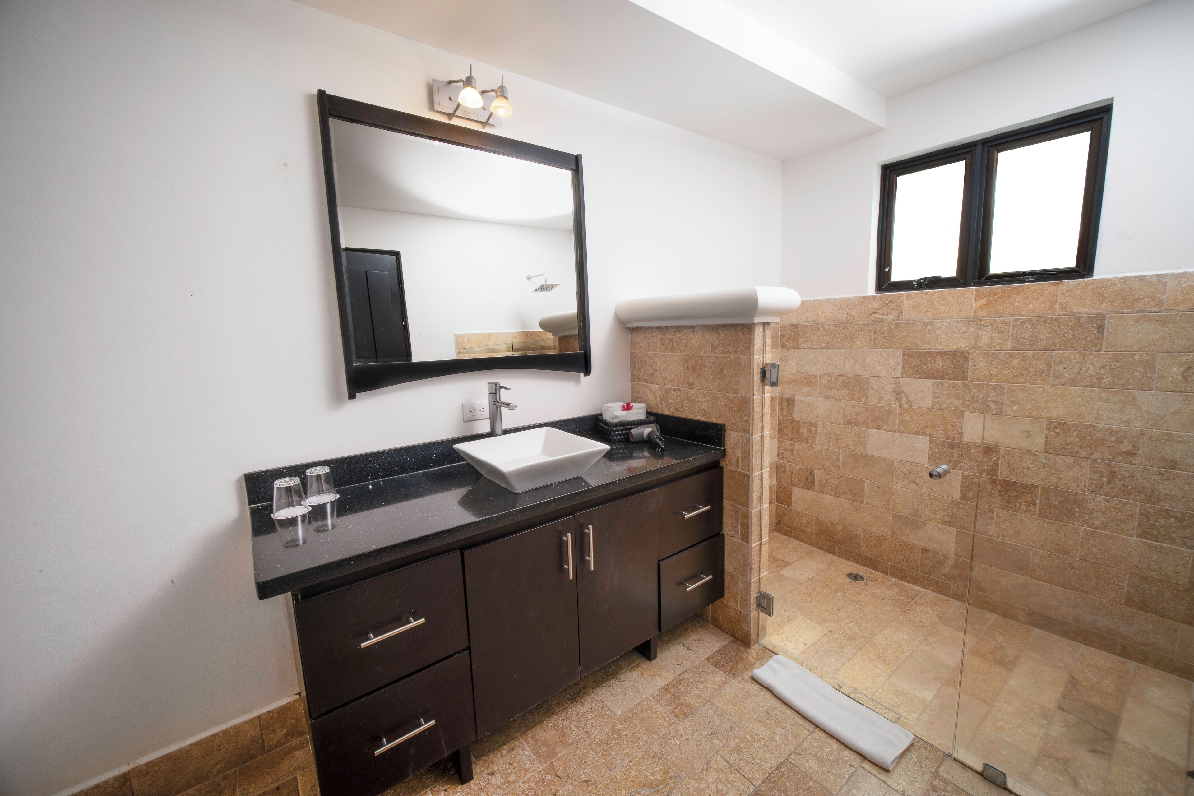Un baño con encimera negra, lavamanos blanco, mueble color café oscuro, espejo y zona de ducha con baldosas beige.